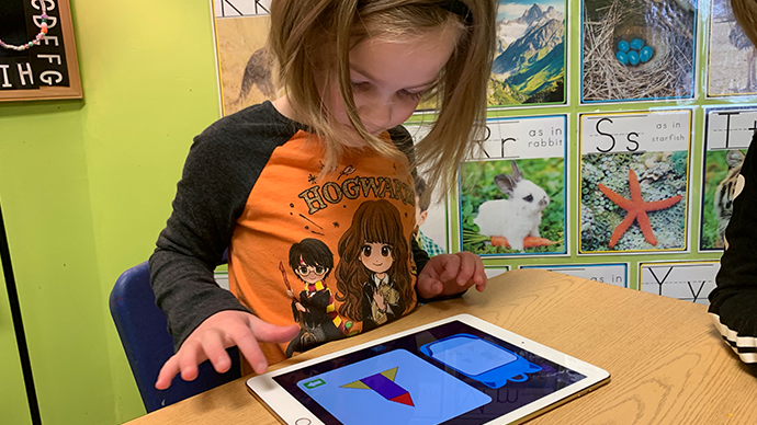 A child is sitting at a laminated wood table and playing an iPad displaying the Shadow Cave app, which has a rocket-shaped puzzle on it made of smaller pieces. 