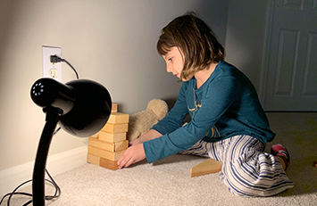 Child stacks blocks to shade a stuffed animal from lamp.
