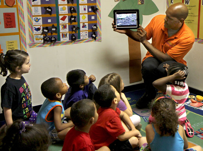 Students watch teacher holding iPad displaying the video.