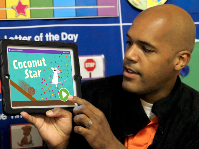 Teacher holds up iPad showing Coconut Star app screenshot.