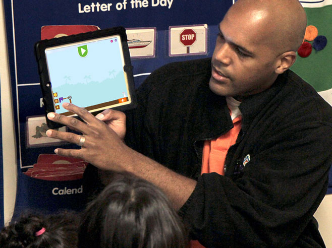 Teacher holds up iPad and points to screen, showing Coconut Star app level screenshot.