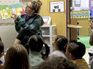 A teacher holds up a book in front of students seated on the floor.