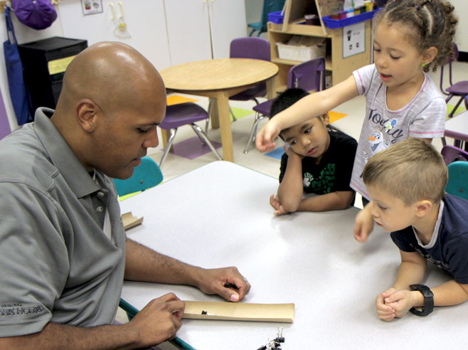 Three students watch as teacher shows them a binder clip, not sliding on a half a cardboard tube.