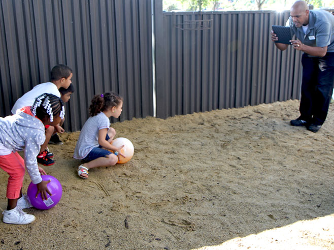 Students in a sandy playground, prepare to roll large plastic balls. A teacher is recording them with an iPad.