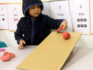 A student holds one piece of flat red play-dough, while rolling a ball of red play-dough down a cardboard ramp.