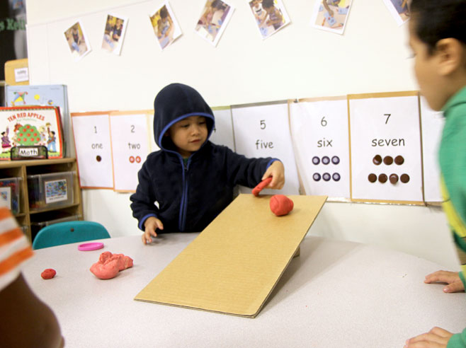 A student holds one piece of red play-dough, while rolling a ball of red play-dough down a cardboard ramp.