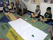 A cardboard ramp leads to a long piece of paper with sticky notes marking distances. A toy car has just come down the ramp and is resting towards the end of the paper.