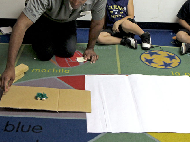 A teacher rolls a toy car down a gently sloping cardboard ramp, which leads to a piece of paper on the floor.