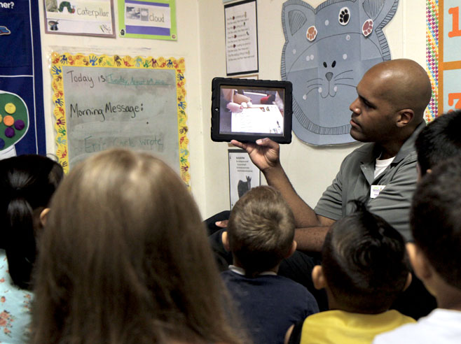A teacher holds up an iPad to show a class of students a video.
