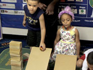 Two students kneel behind cardboard ramps. One student is about to let a round object roll down the steeper of the ramps.