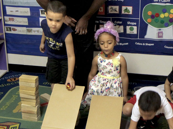 Two students kneel behind cardboard ramps. One student is about to let a round object roll down the steeper of the ramps.
