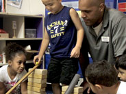 Students and a teacher gather round as a boy prepares to roll a marble down a wooden ruler propped up by wooden blocks.