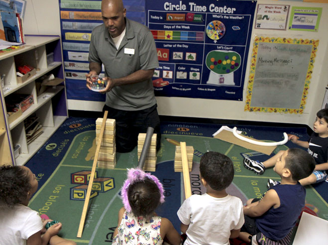 A teacher kneels behind three different ramp set ups, each supported by wooden blocks. He holds a jar filled with marbles.
