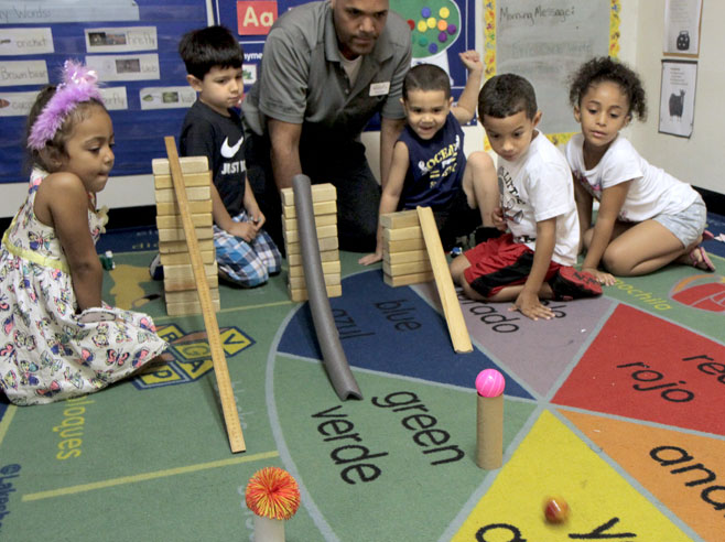 Students and a teacher kneel behind three different ramp set ups. Cardboard tubes with objects balanced on top have been placed, as obstacles, on the floor beyond the ramps. A marble has just been released down one of the ramps.