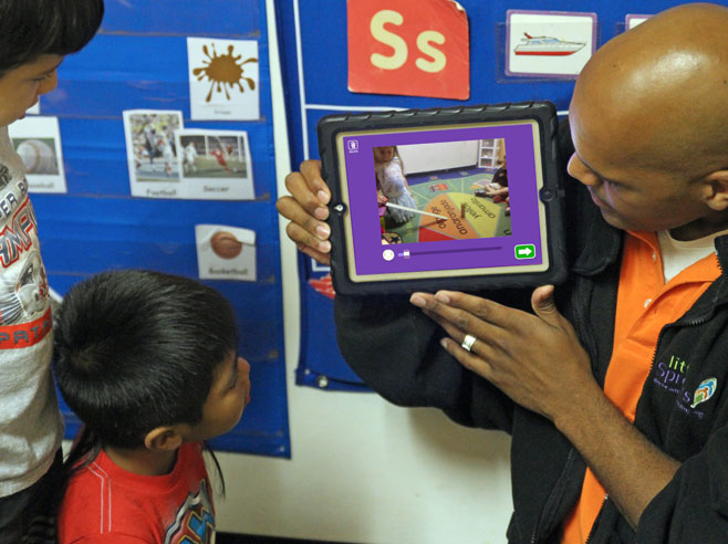 Teacher holds up iPad as students look on.