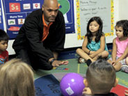 A teacher leans forward, during circle time, to push a purple plastic ball across the rug.