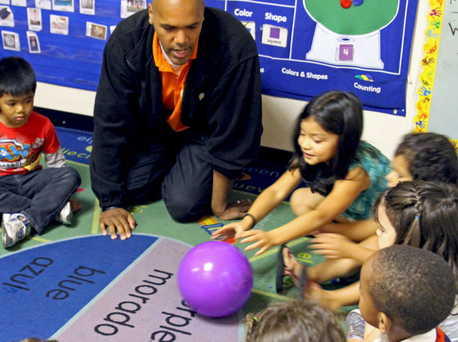 Students and teacher sit in circle time. One student reaches out to push a purple plastic ball across the rug.