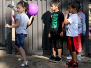 Students stand against a fence. One girl holds a pink ball, raised as if ready to throw.