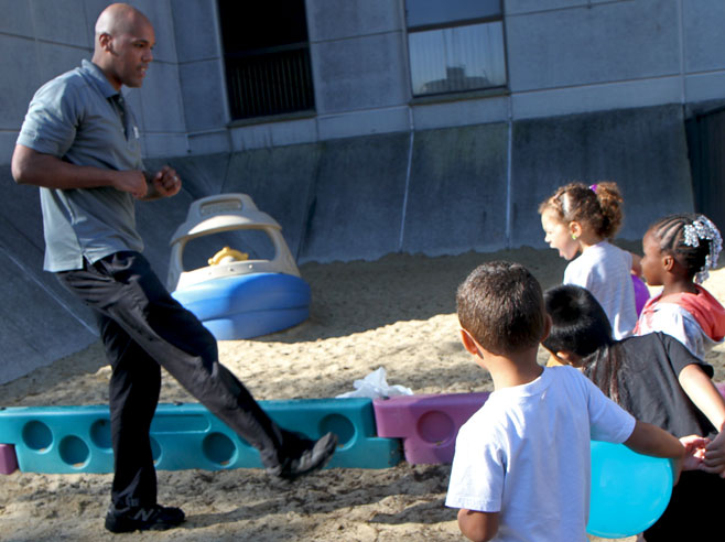In a sandy playground, teacher makes kicking motion with foot as students look on.