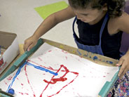 A student tilts a tray with a piece of paper, a marble, and some red and blue paint. The marble is rolling and creating patterns with the paint.