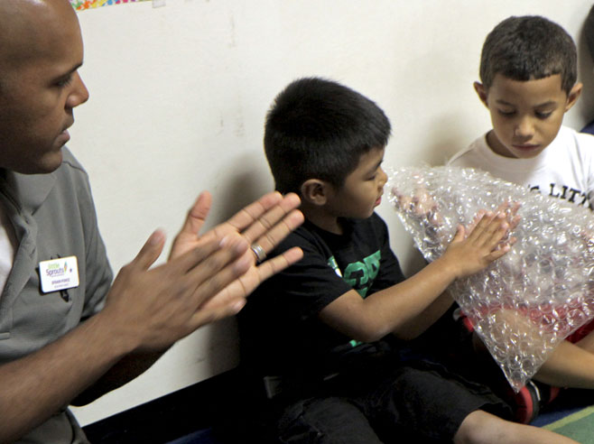 A teacher motions with his hands, as two students feel a piece of bubble wrap.