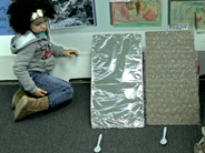 A student sits on the floor next to two cardboard ramps leaning against the wall. One ramp is covered in tin-foil and the other is covered in bubble wrap.