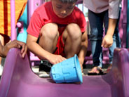 A student crouches at the top of a slide and prepares to roll a blue plastic bucket down the slide.