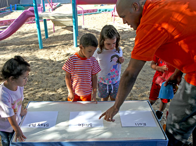 In a sandy playground, a table has been set up with three pieces of paper, one that reads 'roll', one 'slide', and one 'stay put'. A teacher points at the piece of paper that reads 'slide' as students look on.