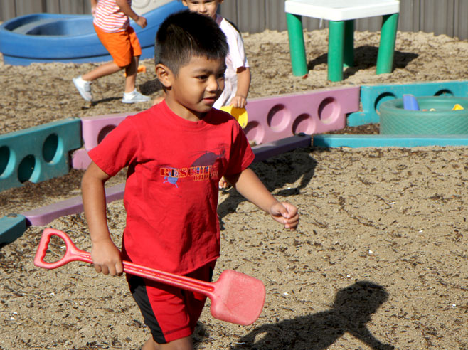A student carries a toy red plastic shovel across a sandy playground.