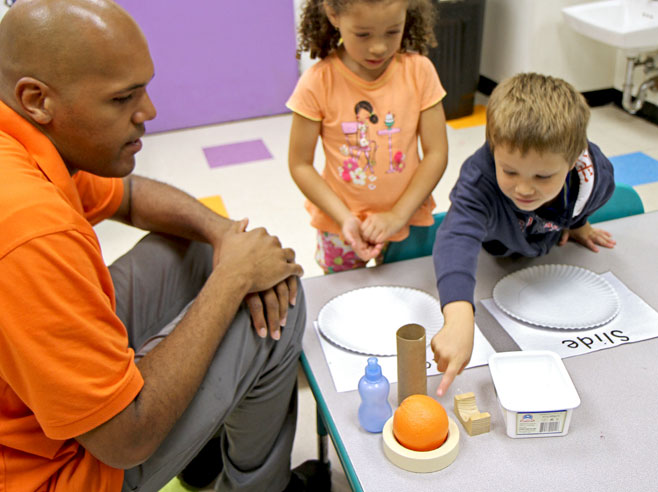 At a table two students and a teacher gather round an assortment of objects. One student points at one of the objects, an orange.