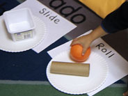A student’s hand places an orange next to a cardboard tube, onto a paper plate with the word 'Roll' written next to it. Close by, a plastic carton sits on a paper plate with the word 'Slide' next to it.