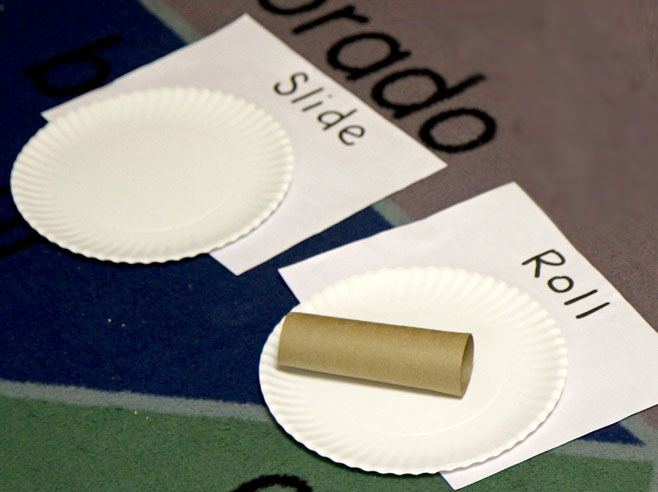A cardboard tube sits on a paper plate with the word 'Roll' written next to it. Close by, is another paper plate, with the word 'Slide' next to it.