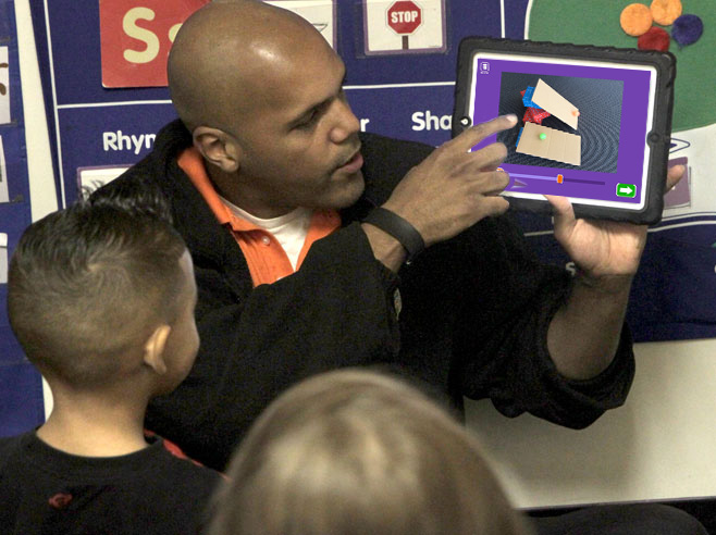 A teacher holds an iPad up and points to the screen showing two balls rolling down two differently sloping cardboard ramps. Students look on.