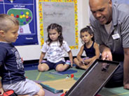 Students look on, as a teacher holds a baking tray at an angle from the floor, and slides a toy plastic turtle down the tray.