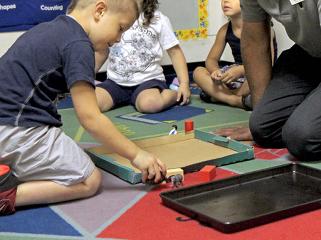 A large box lid and a baking tray lie on the floor, surrounded by several small toy objects. Students and a teacher are kneeling, and one student picks up a toy.