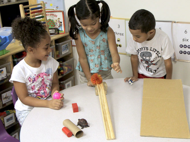 Three students gather at a table with some ramps made from different materials. They hold toys, and one sends a pompom ball down a ramp.