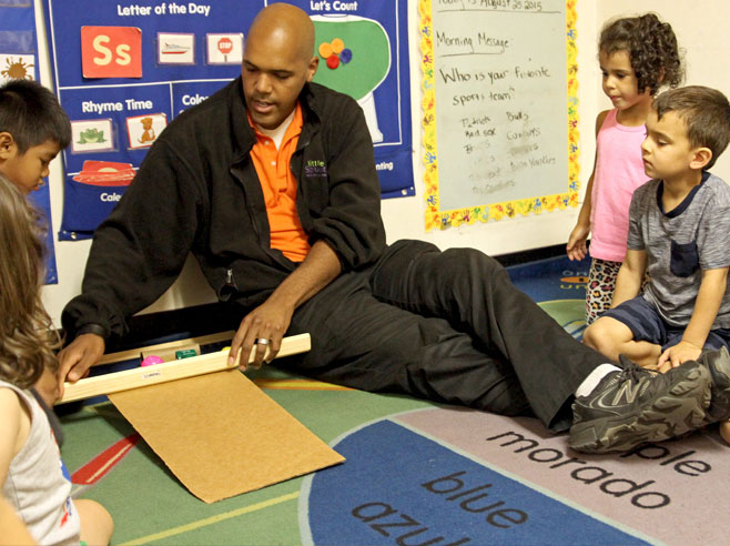 A teacher sits on the floor next to a cardboard ramp. He holds a wooden stick in front of a pink plastic ball and a toy car, to keep them from going down the ramp.