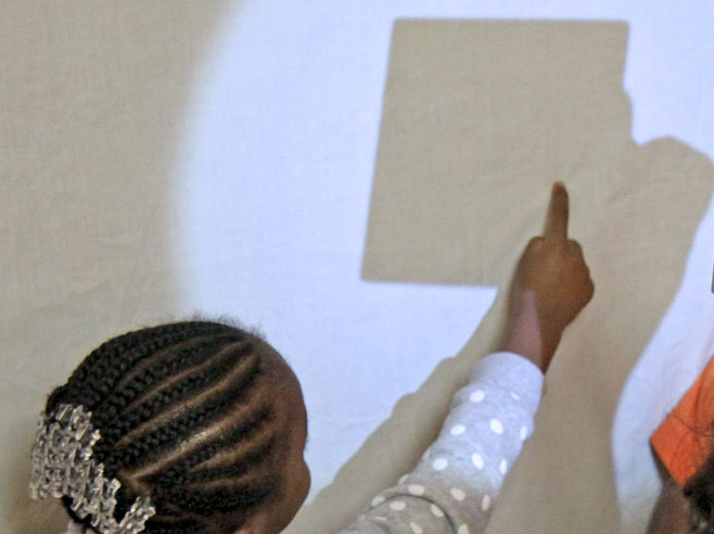 There is a shadow, on a spot lit wall, of someone's arm holding a wooden block. A student points at the shadow.