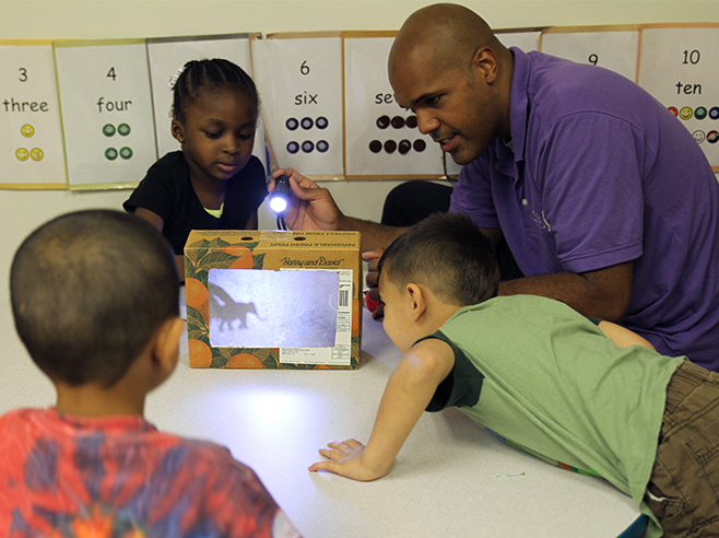 Teacher and student shining flashlight at a shadow box theater. Two students watch the shadows.