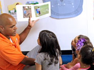 A teacher holds a book up to his students. The book is open to show some photos.