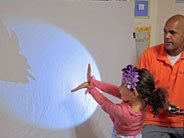 A student makes shadows with her hands, against a spot lit wall, as a teacher looks on.