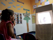 Two students sit in chairs and watch a shadow puppet show that is set up in front of them.