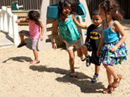Four students are dancing in a sandy playground. It is sunny, and their shadows are visible.