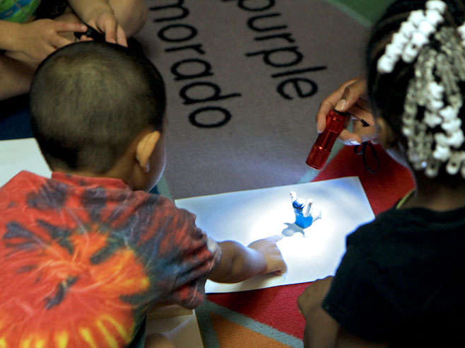 A flashlight shines on a small toy that is placed on a piece of white paper on the floor. Students look on, and one points at the shadow created by the toy.