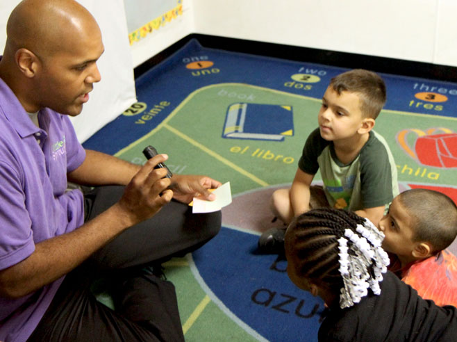 A teacher sits on the floor talking to three students. He holds a flashlight in one hand, and some square pieces of paper in the other.