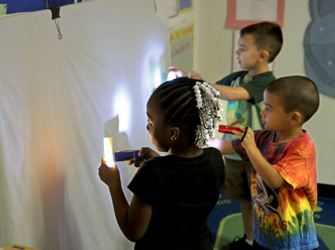 Three students stand at a wall holding flashlights and pieces of square paper to create shadows on the wall.