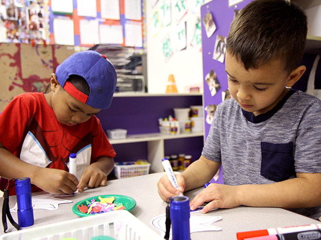 Two students sit at a table. They are both coloring with markers. Collage materials can be seen on the table.