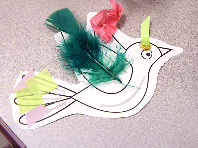 A 2-D cut-out of a bird lies on a table. Different collage elements have been glued to the bird, such as feathers and tissue paper.