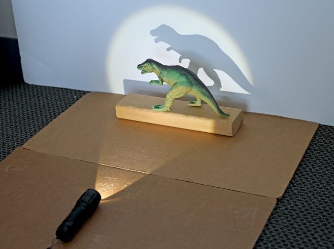 A toy dinosaur stands on a wooden block, against a white wall. A flashlight lies on the floor, pointed at the dinosaur, and creates a shadow of the dinosaur on the wall behind.