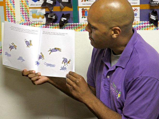 A teacher holds a book up, open to a page that shows illustrations of rabbits jumping. The shadows of the rabbits are visible.